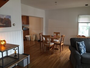 A cozy living room with a dark sofa and a wooden dining table with chairs. A kitchen is visible in the background with wooden cabinets and a window.