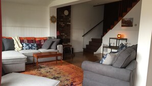 A living room with a sofa set, a coffee table, and a patterned rug. A staircase is visible in the background leading to another level.