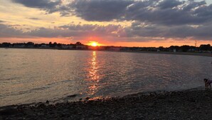 A sunset over a calm river with a rocky shoreline and a distant cityscape.