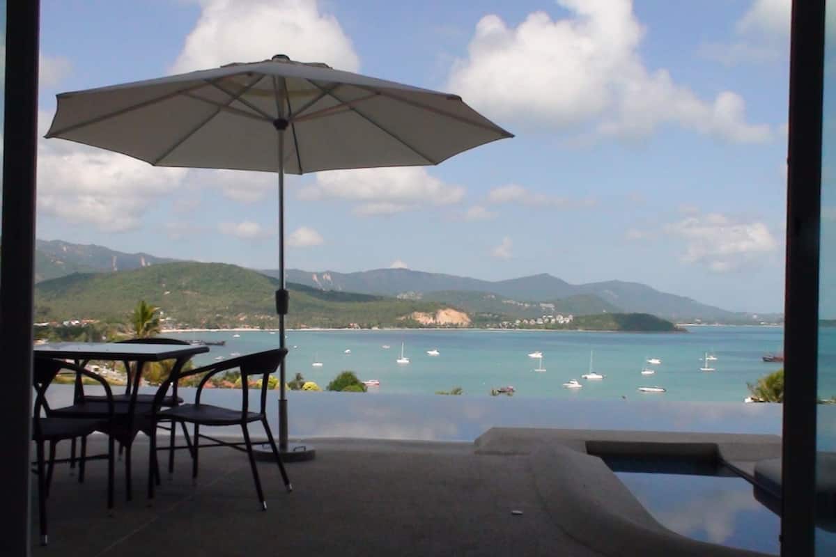 An infinity pool with a view of the sea and mountains. There is a table and chairs under an umbrella.