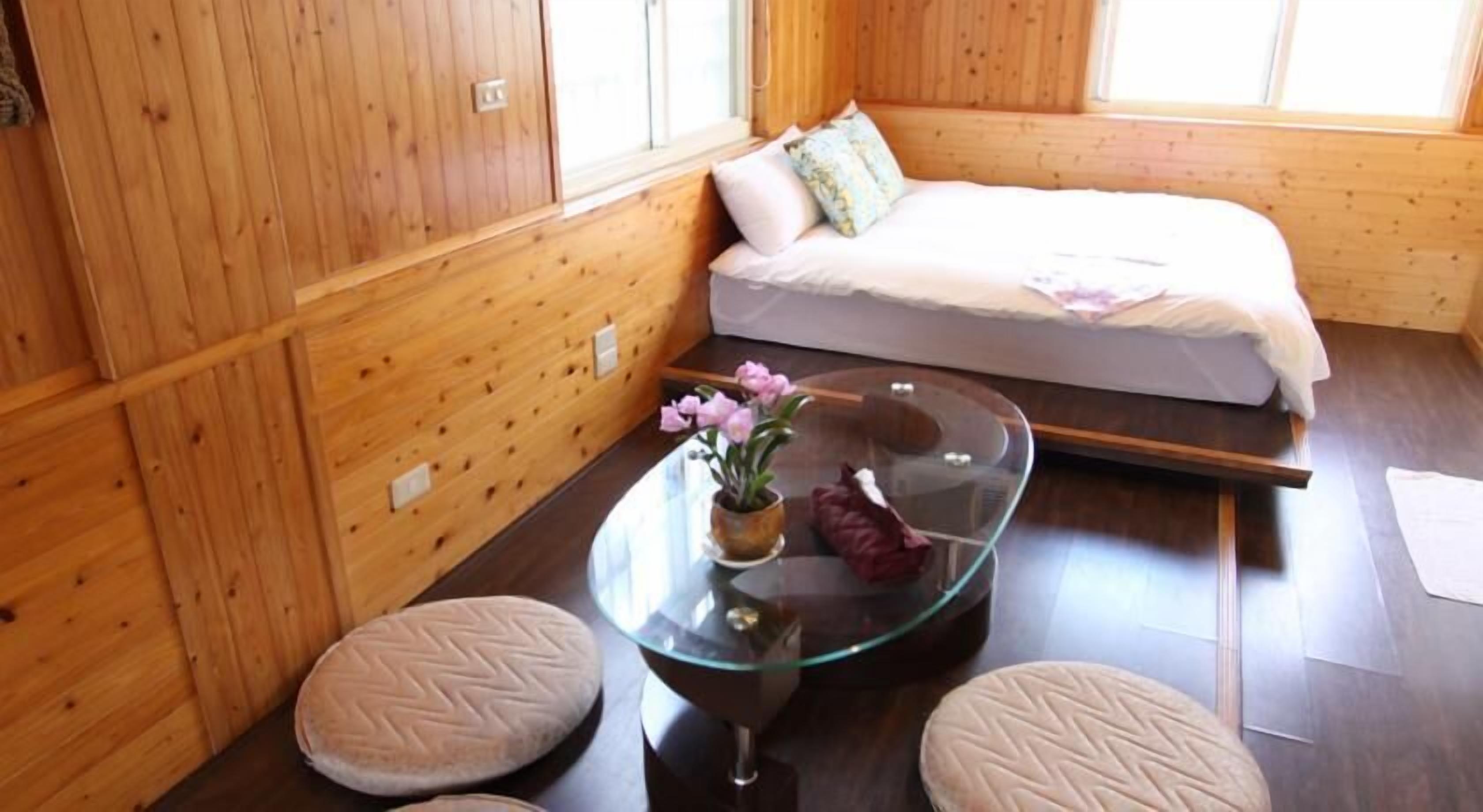 A wooden cabin room with a bed, a glass table with a flower, and two ottomans.