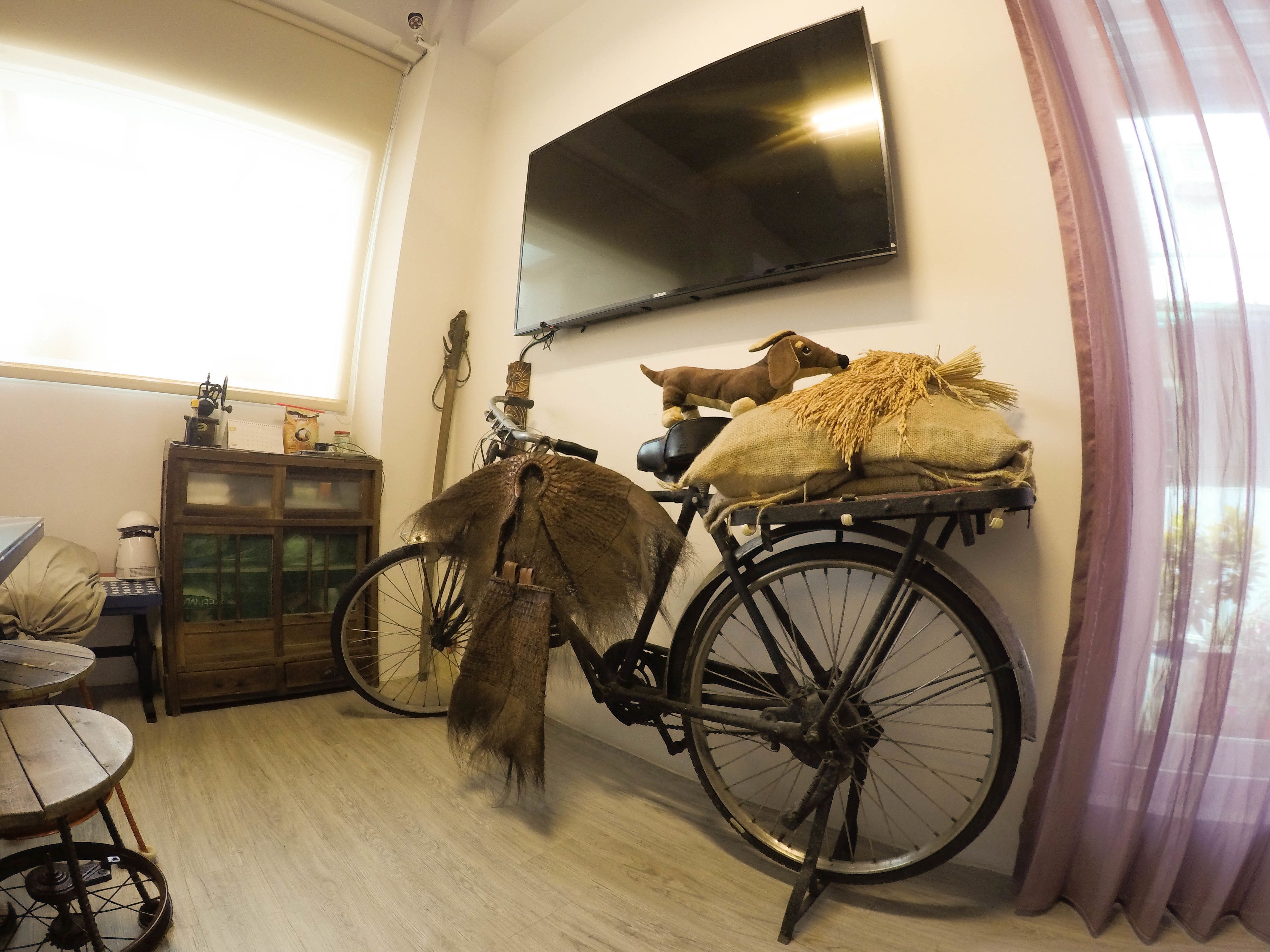 A room with a wooden floor, a bicycle with a basket, a television mounted on the wall, and a window with curtains.