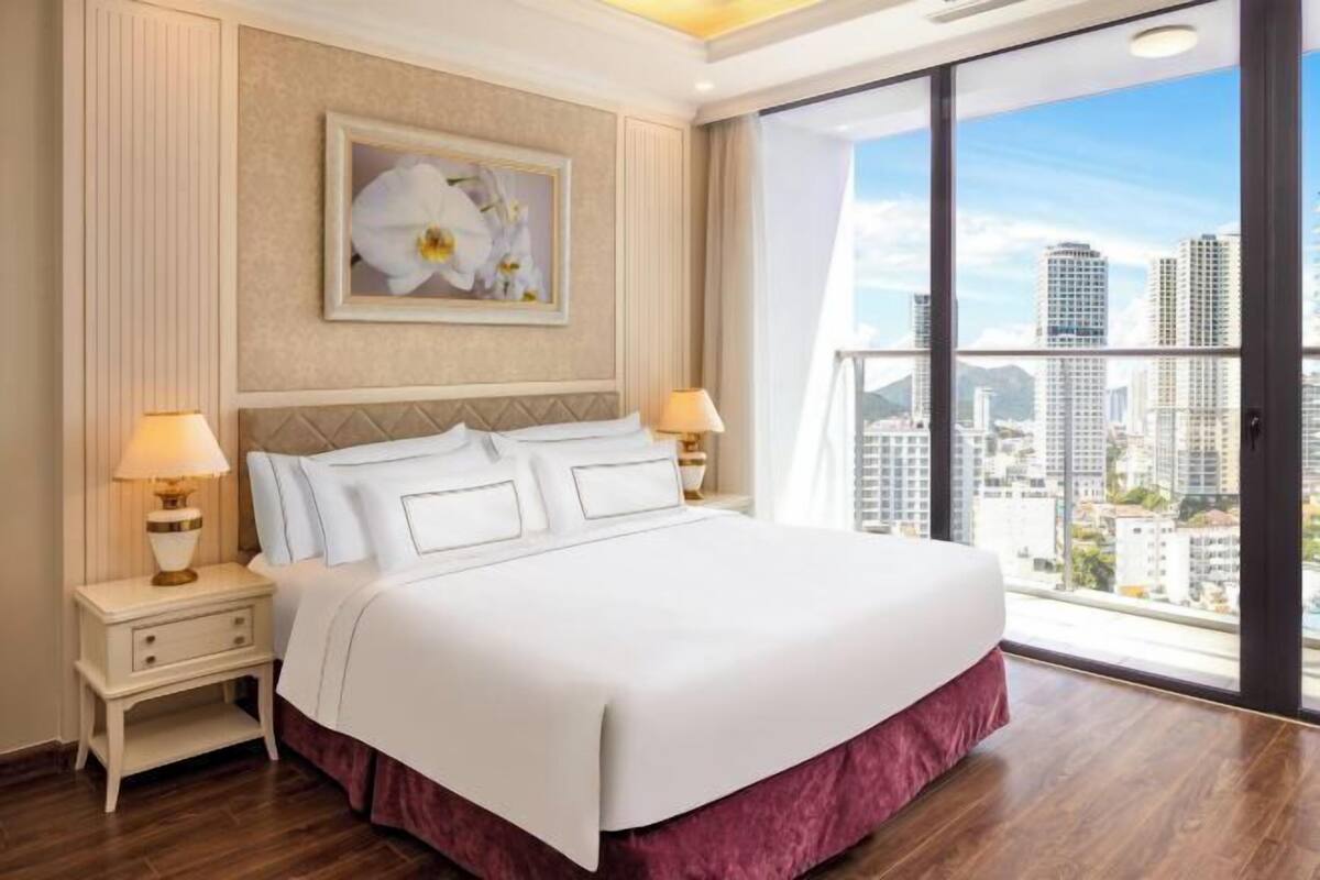 Quarto de hotel com uma cama grande, duas mesas de cabeceira com luminárias, um quadro na parede e vista para a cidade através de janelas amplas.
