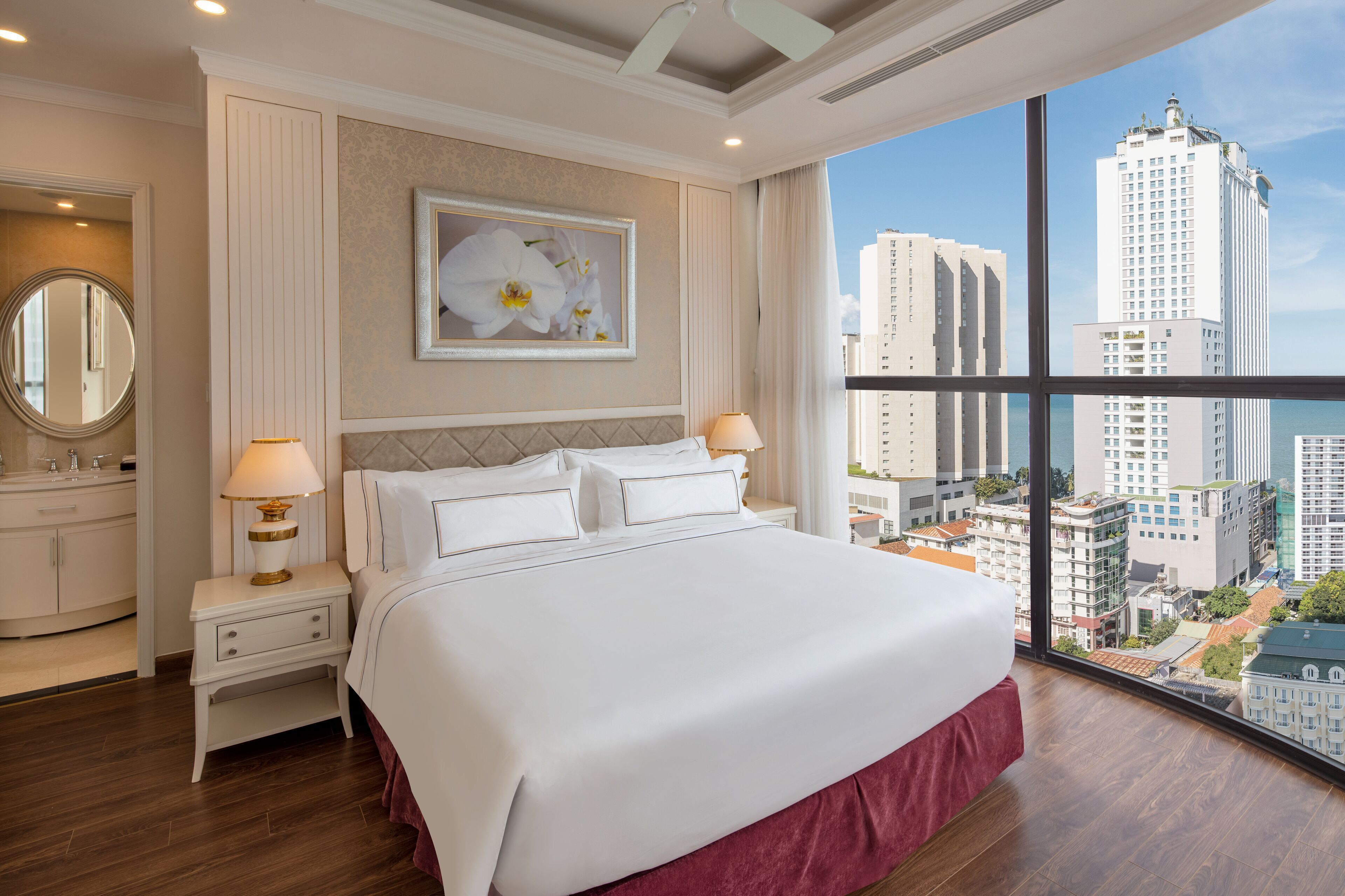 Une chambre d’hôtel avec un grand lit, deux tables de chevet avec des lampes, un grand miroir rond et une vue sur de hauts immeubles.