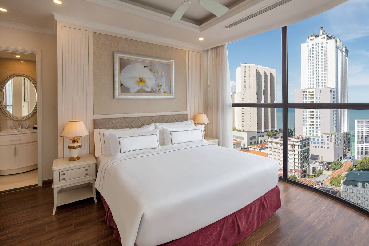 Quarto de hotel com uma cama grande, duas mesas de cabeceira com abajures, um espelho redondo e vista para edifícios altos.
