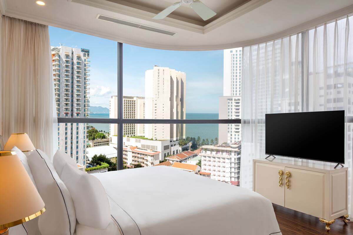 Quarto de hotel com uma cama grande, uma televisão sobre um móvel e vista para edifícios altos e o mar.