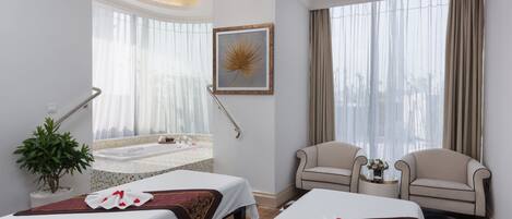 Habitación de hotel con dos camas, un espacio de descanso, una mesita con un florero y un ventanal grande con cortinas.
