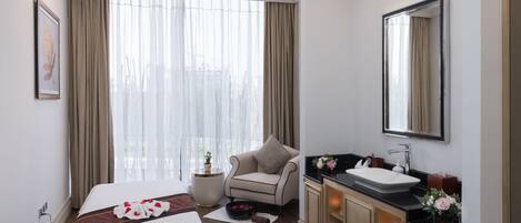 A hotel room with a bed, a sitting area with a chair, a vanity with a mirror, and a window with curtains.
