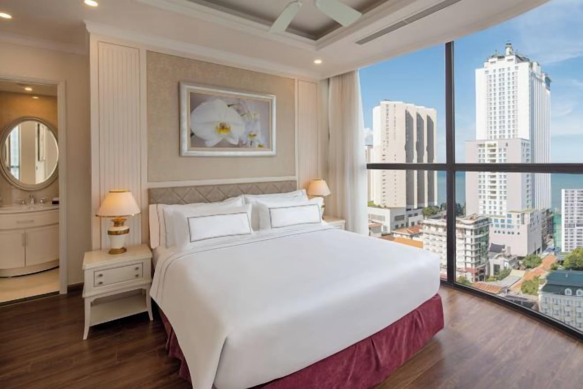 Quarto de hotel com uma cama grande, mesa de cabeceira com abajur, espelho e vista para a cidade.