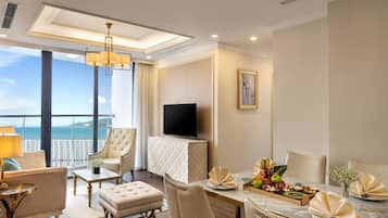 A hotel room with a balcony, a TV, a dining table set with fruit and flowers, and a large window with a view of the sea.