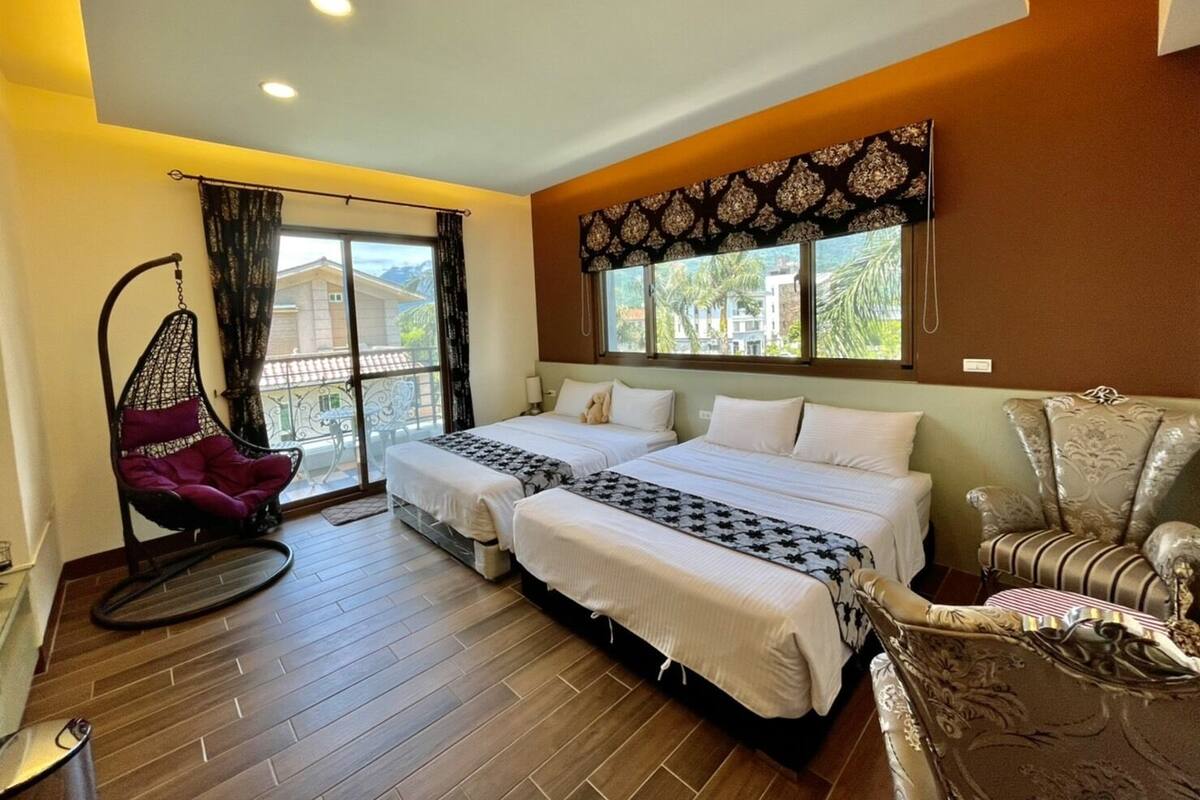 Habitación de hotel con dos camas, una silla colgante, un sofá y un ventanal con vistas a edificios y árboles.