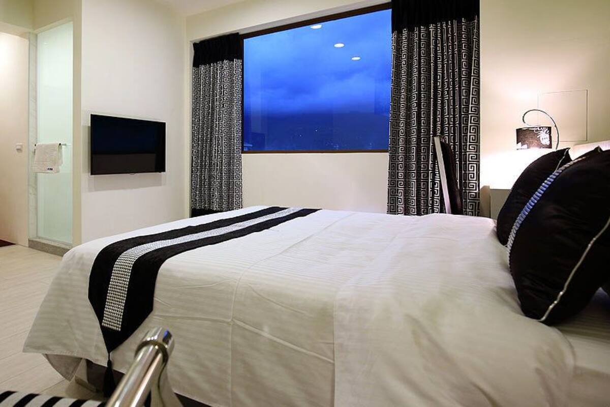 Habitación de hotel con cama, televisor montado en la pared, ventana con cortinas y una mesita de noche con lámpara.