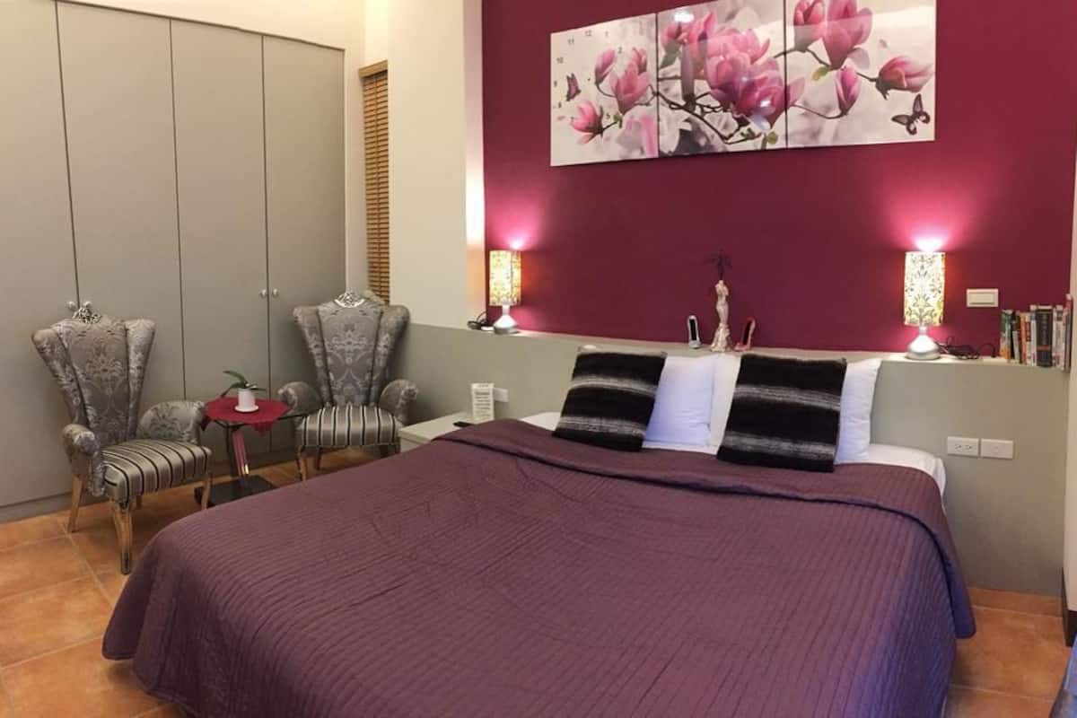 Un dormitorio con una cama grande, dos sillas, una mesita de noche y una estantería en la pared con libros y objetos decorativos.
