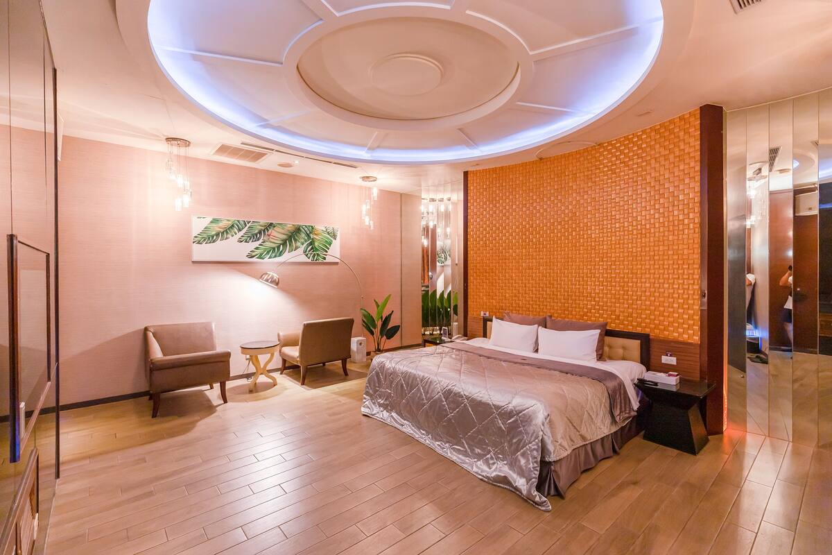 A modern hotel room with a large bed, a seating area with a chair and a small table, a wall-mounted artwork, and a unique orange brick wall.