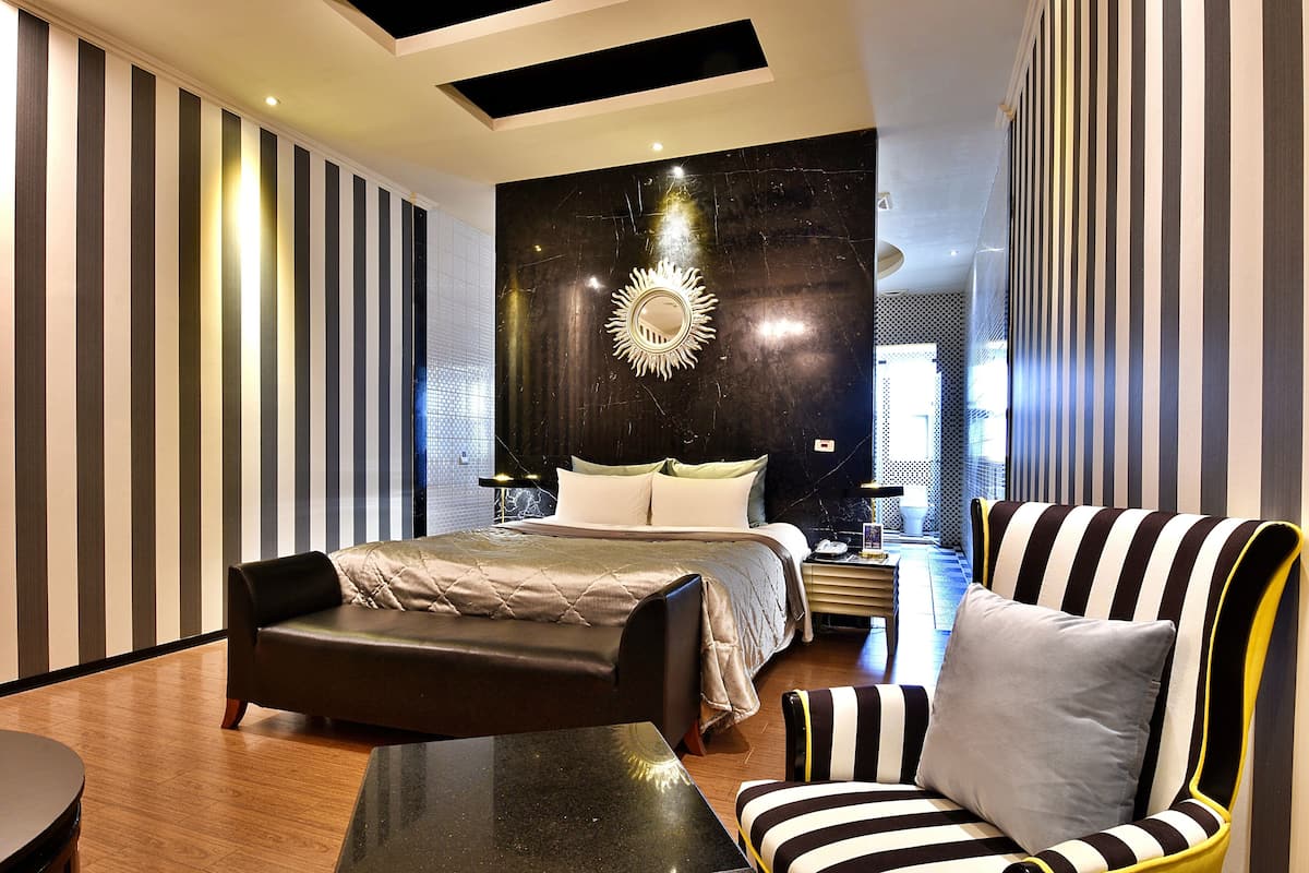 A modern hotel room with a large bed, a black leather sofa, a striped armchair, and a unique wall mirror.