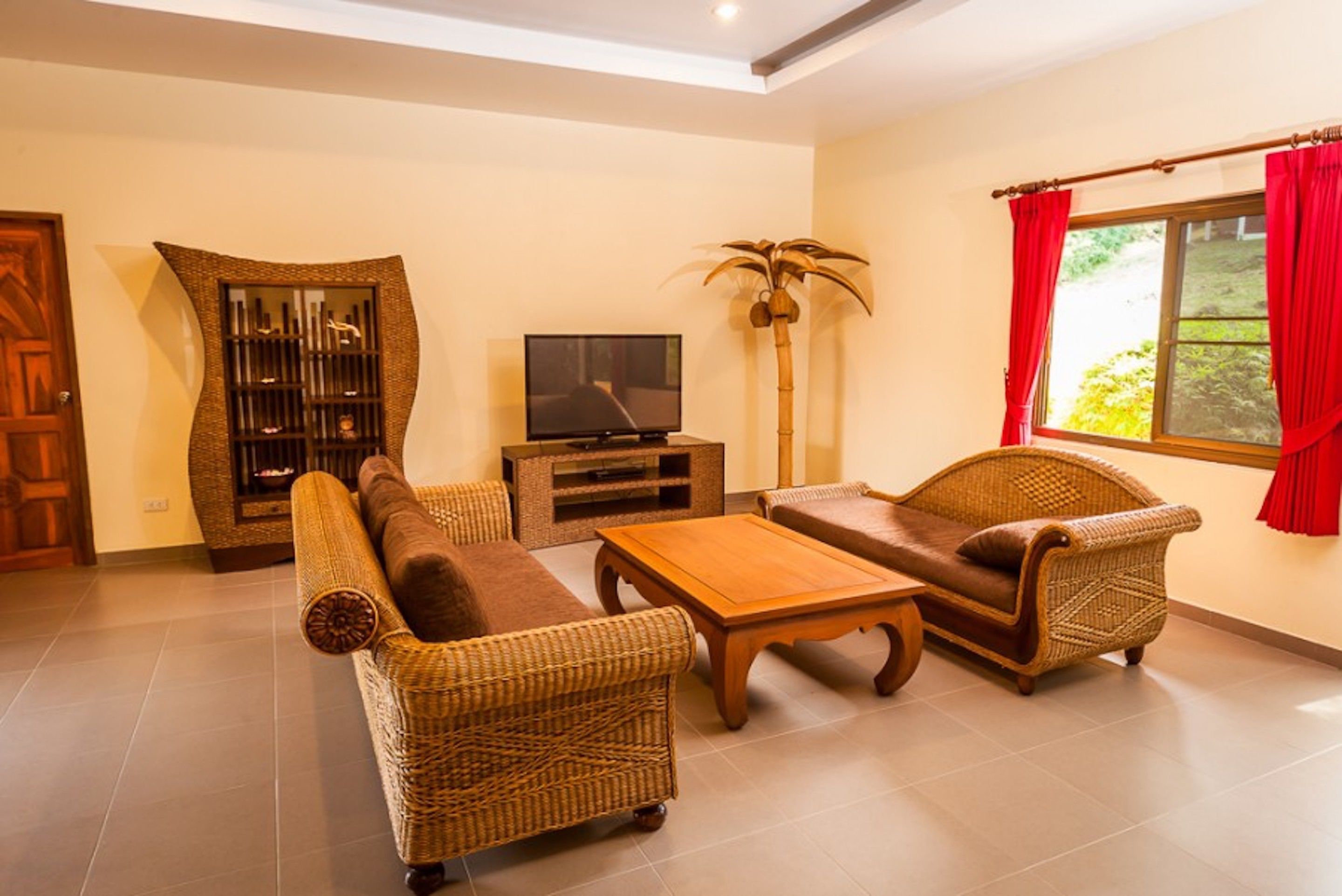 A living room with wicker furniture, a television, and a large window with red curtains.