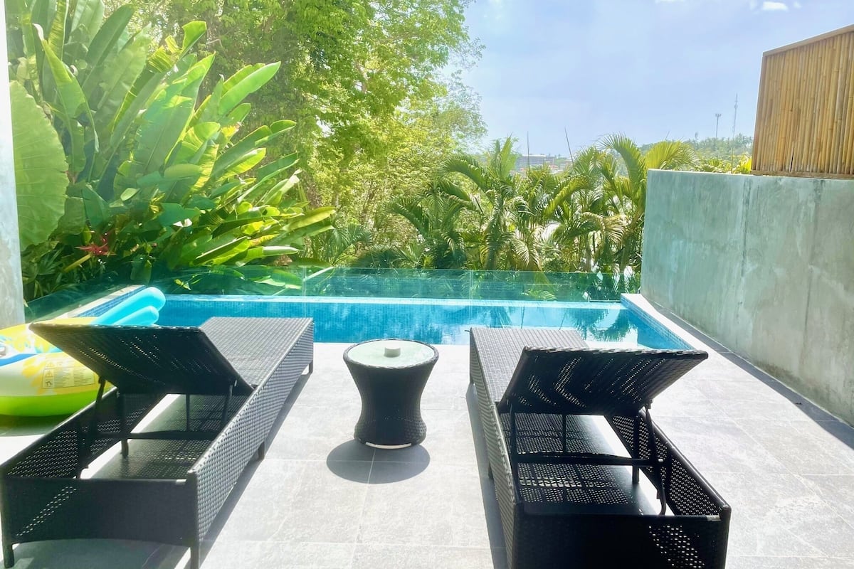 Un área de piscina en la azotea con dos sillones de descanso y una mesita pequeña, rodeada de abundante follaje y un cielo azul despejado.