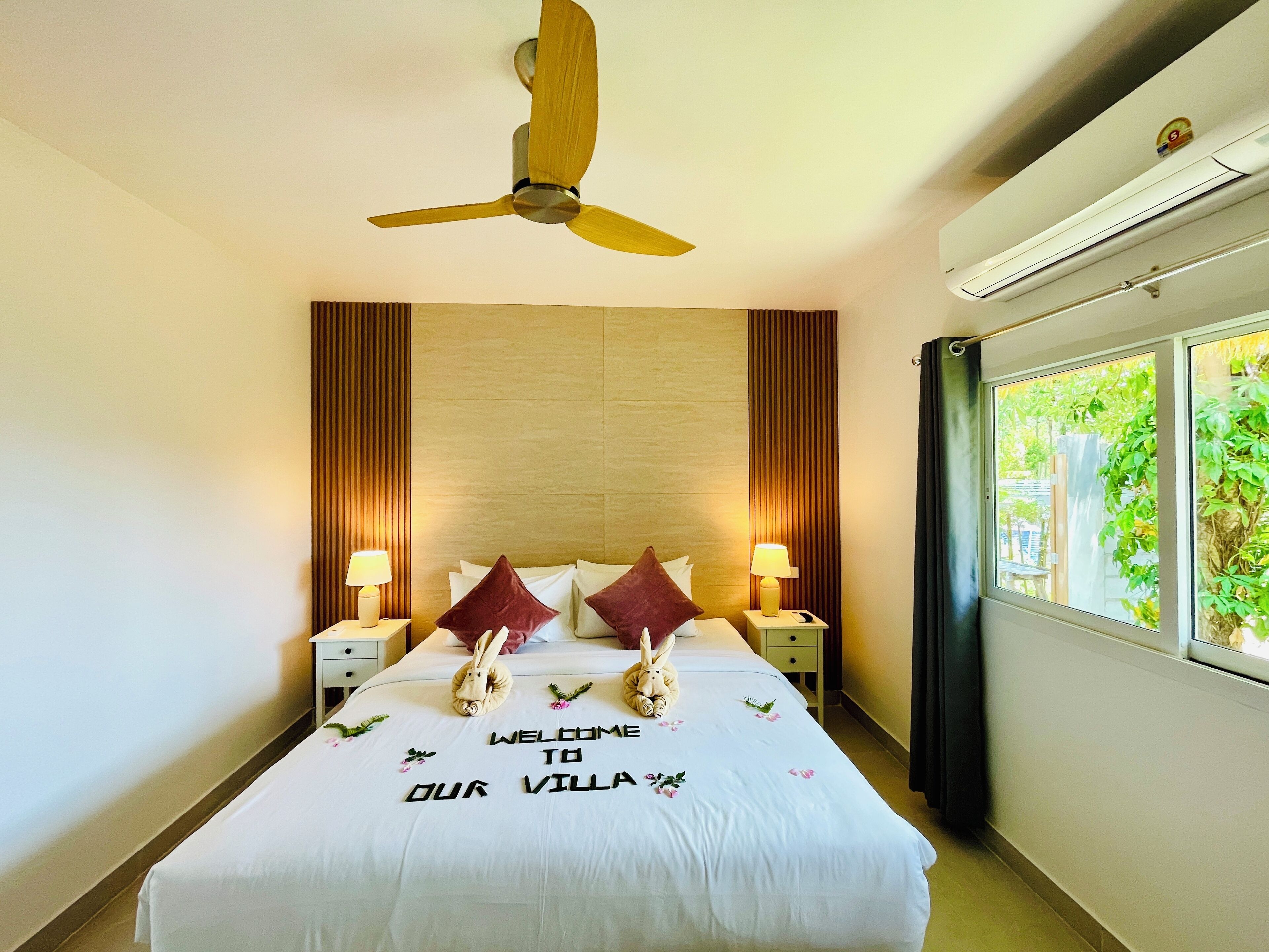 Habitación de hotel con cama, dos mesitas de noche, ventilador de techo, ventana con vistas y aire acondicionado de pared.
