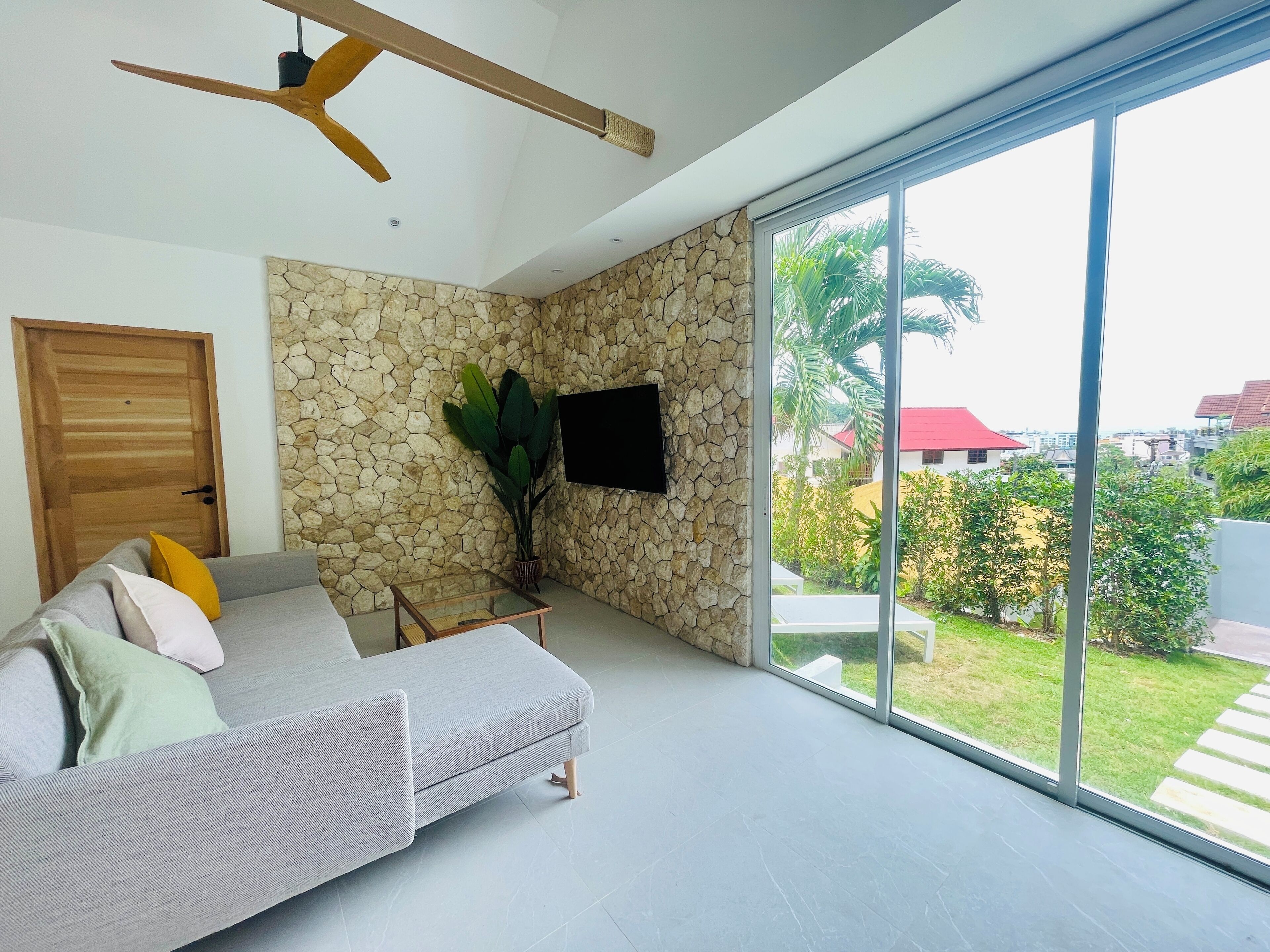 A modern living room with a stone wall, a grey sofa, a wooden ceiling fan, and a large sliding glass door leading to a garden.