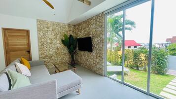 Uma sala de estar moderna com parede de pedra, sofá cinza, ventoinha de teto de madeira e uma grande porta de vidro deslizante que dá para um jardim.