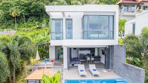 Ein modernes, zweistöckiges Haus mit Pool, Sitzgelegenheiten im Freien und Küchenbereich.