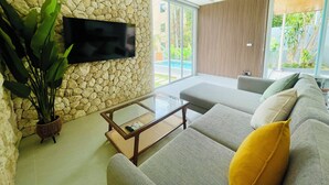 A modern living room with a stone wall, a flat-screen TV, a glass coffee table, and a large sofa with cushions. - Baan Saint Tropez Villas Kata Beach (Karon)