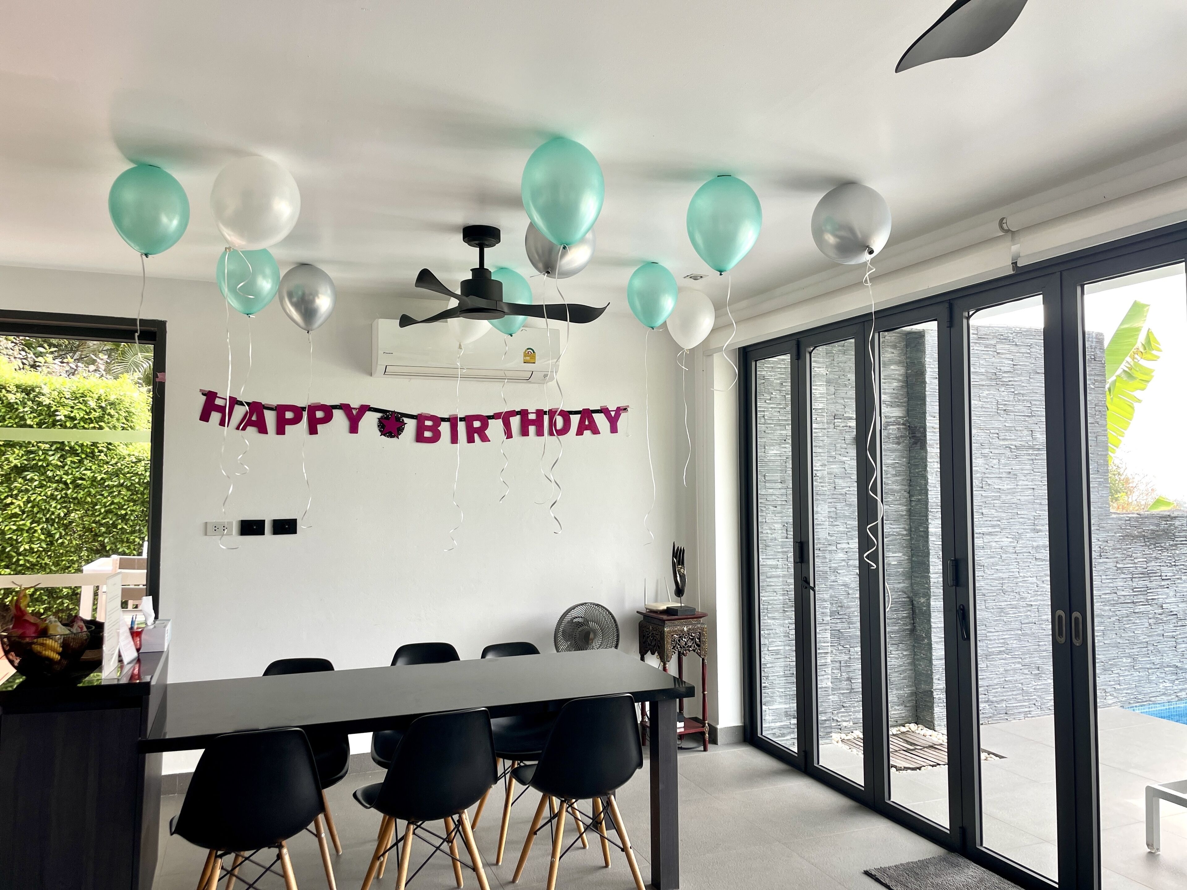 Een moderne eethoek met een zwarte tafel en stoelen, een plafondventilator en een 'Happy Birthday' spandoek.