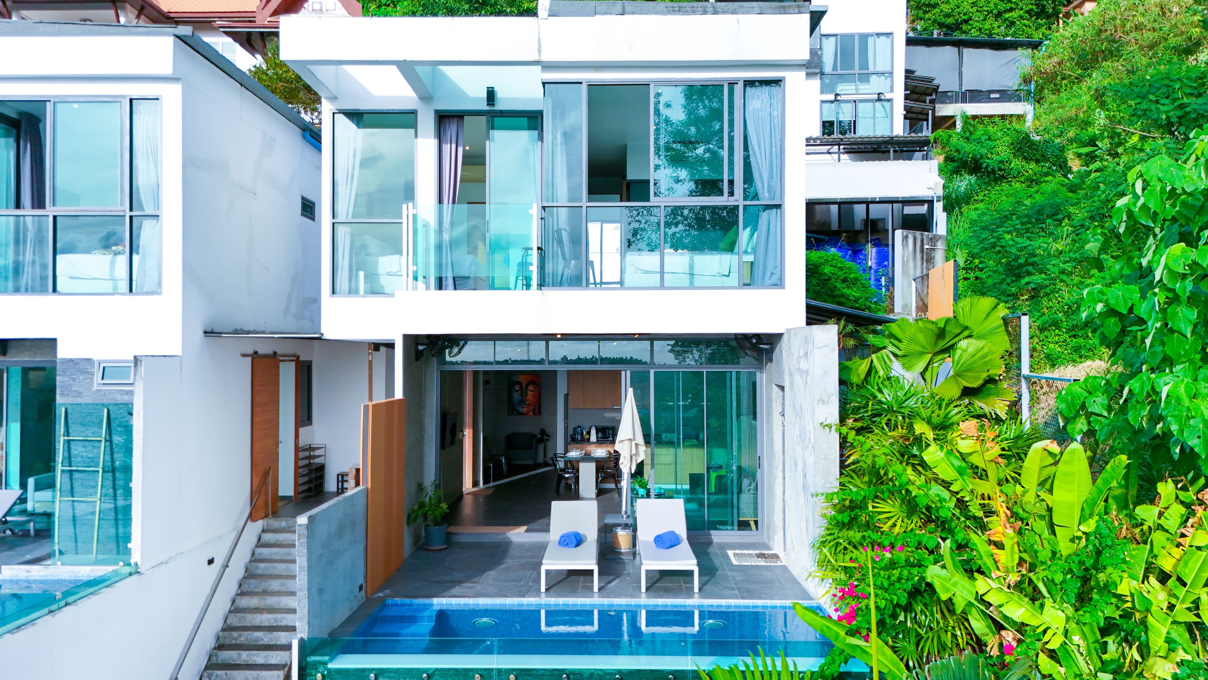 Una casa moderna de varios pisos con fachada de vidrio, piscina y balcón con sillones de descanso.
