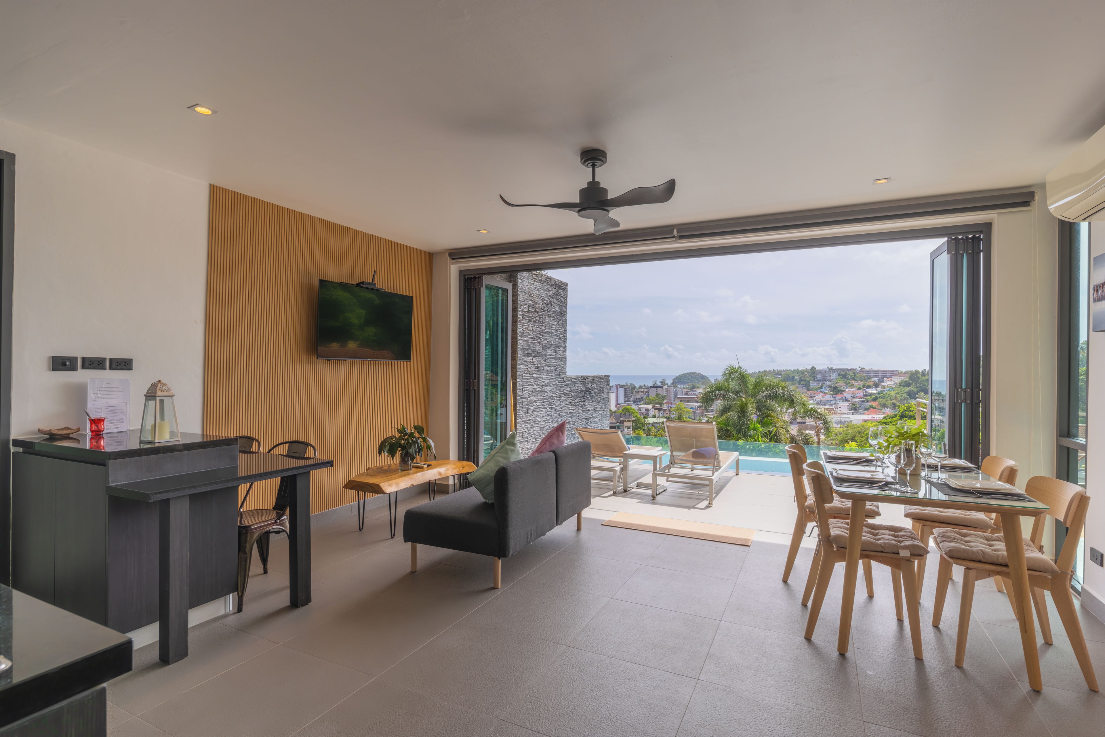 A modern living room with a flat-screen TV, a dining area with a table set for two, and a balcony with a view of the city.