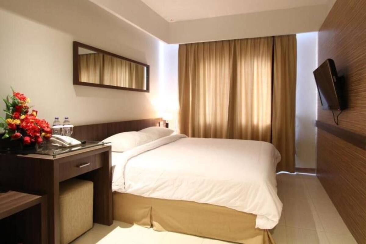 A hotel room with a bed, a desk with a telephone, a mirror, a TV mounted on the wall, and a window with curtains.