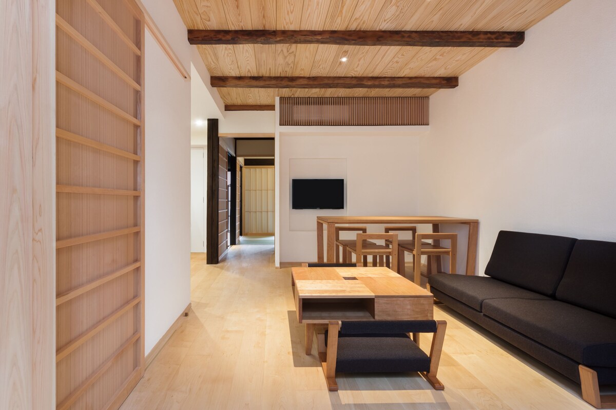 A modern living room with a wooden floor, a black sofa, a wooden coffee table, and a TV mounted on the wall.