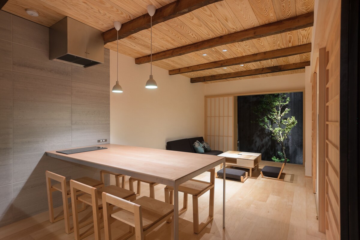 Un interior moderno con una mesa de comedor de madera, sillas, luces colgantes y vistas a la vegetación a través de una puerta corredera.