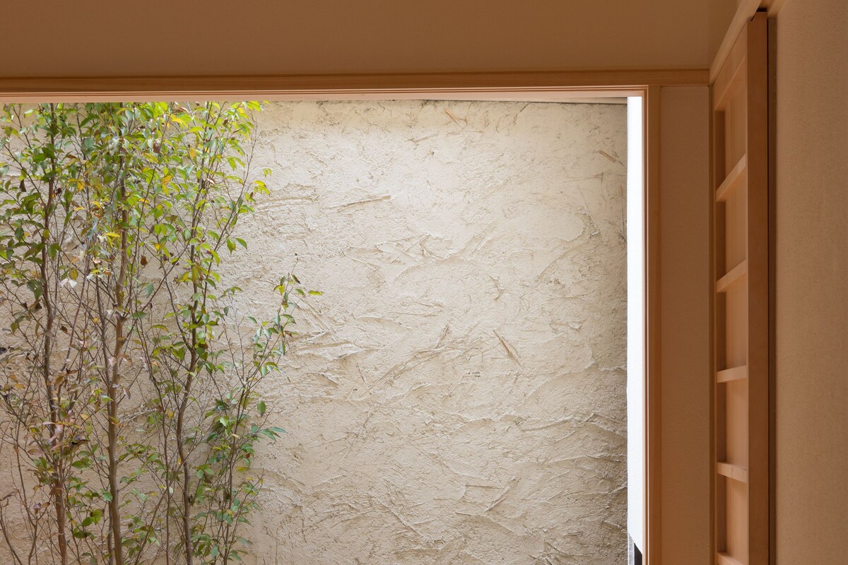 Una habitación con un escritorio de madera, un banco y vistas a una pared con textura y un árbol afuera.