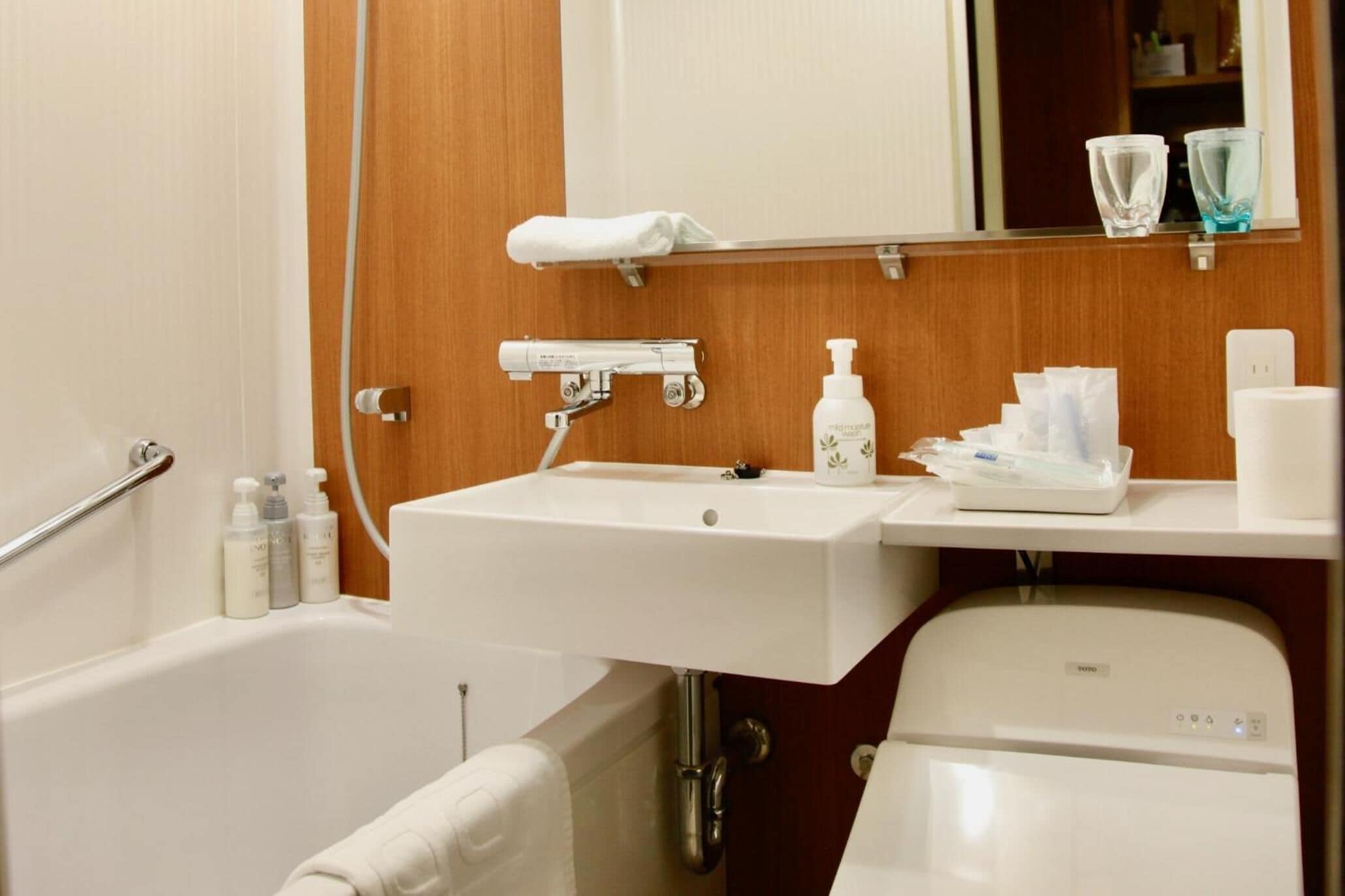 A bathroom with a white sink, mirror, and bathtub. There are toiletries on the sink and a towel rack above the mirror.
