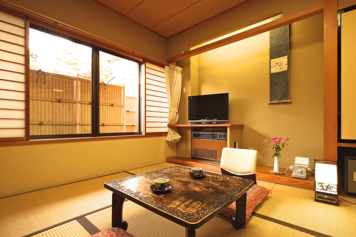 Une pièce de style japonais traditionnel, avec un sol recouvert de tatamis, une petite table sur laquelle sont posées deux tasses, un téléviseur et un petit bureau avec un téléphone et un vase de fleurs.