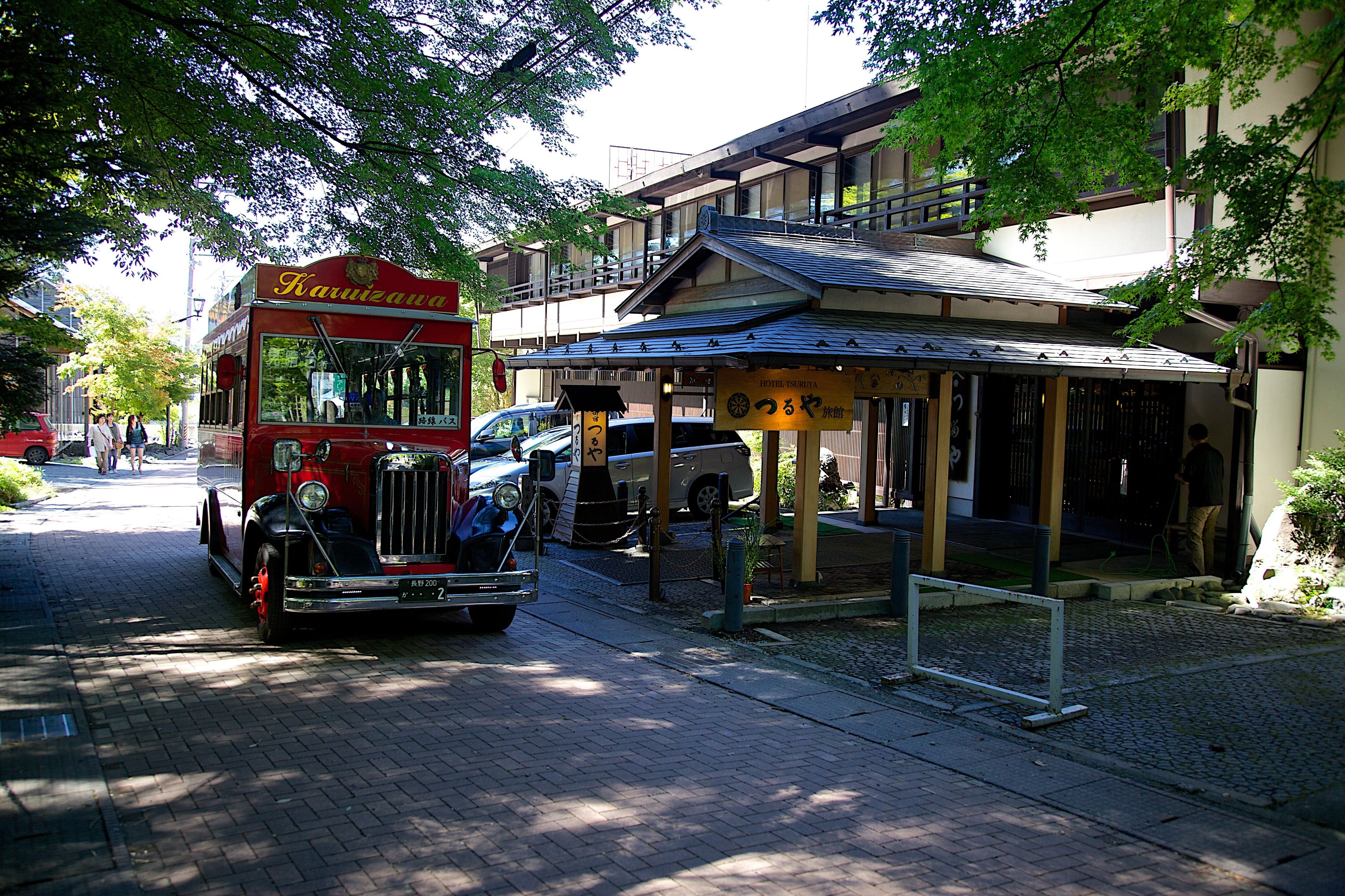 Un autobus rosso d'epoca parcheggiato su una strada acciottolata, un edificio con un ingresso coperto e un'auto nelle vicinanze.
