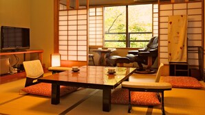 Een traditionele Japanse kamer met tatami vloerbedekking, een lage tafel, stoelen, een televisie en een groot raam met uitzicht op bomen.