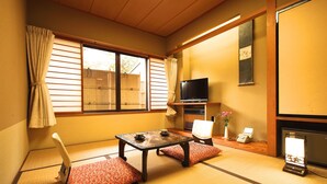 Een traditionele Japanse kamer met tatami vloeren, een lage tafel met kussens, een televisie en een schuifdeur naar een tuin.