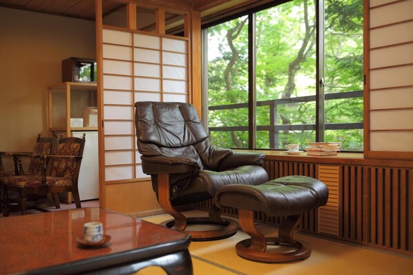 A traditional Japanese room with tatami flooring, a tatami mat table, a leather chair, a wooden cabinet, and a large window with a view of greenery.