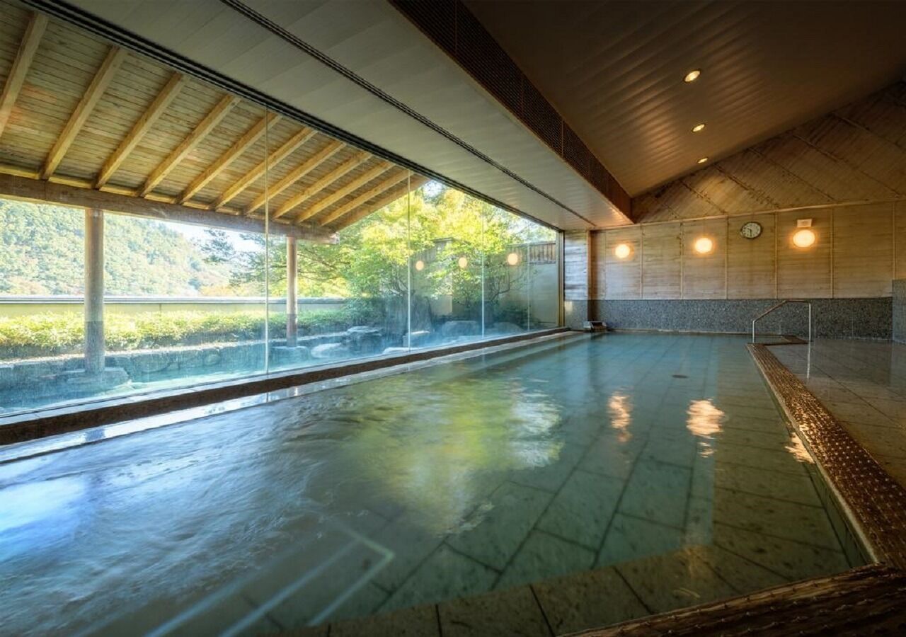 A swimming pool with a glass enclosure, wooden ceiling beams, and a view of greenery outside.