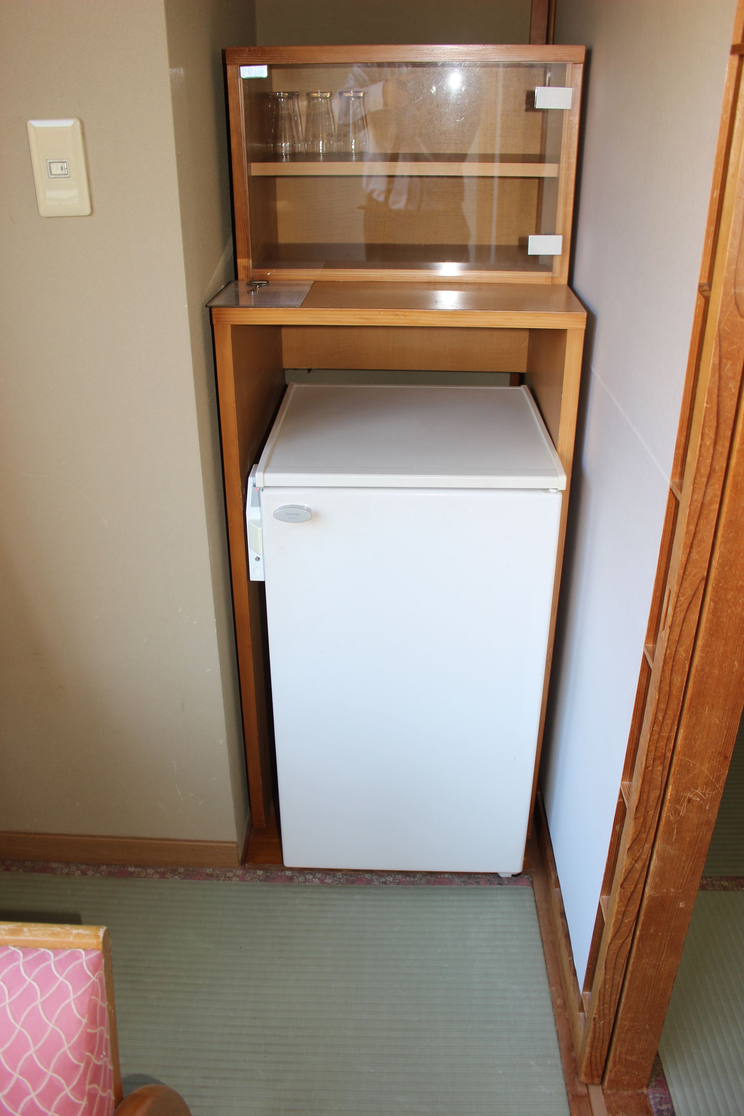 A compact room with a white refrigerator, a wooden cabinet with glass doors, and a small wooden table.