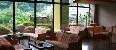 A spacious living area with multiple sofas and a coffee table, large windows offering a view of greenery and buildings outside.