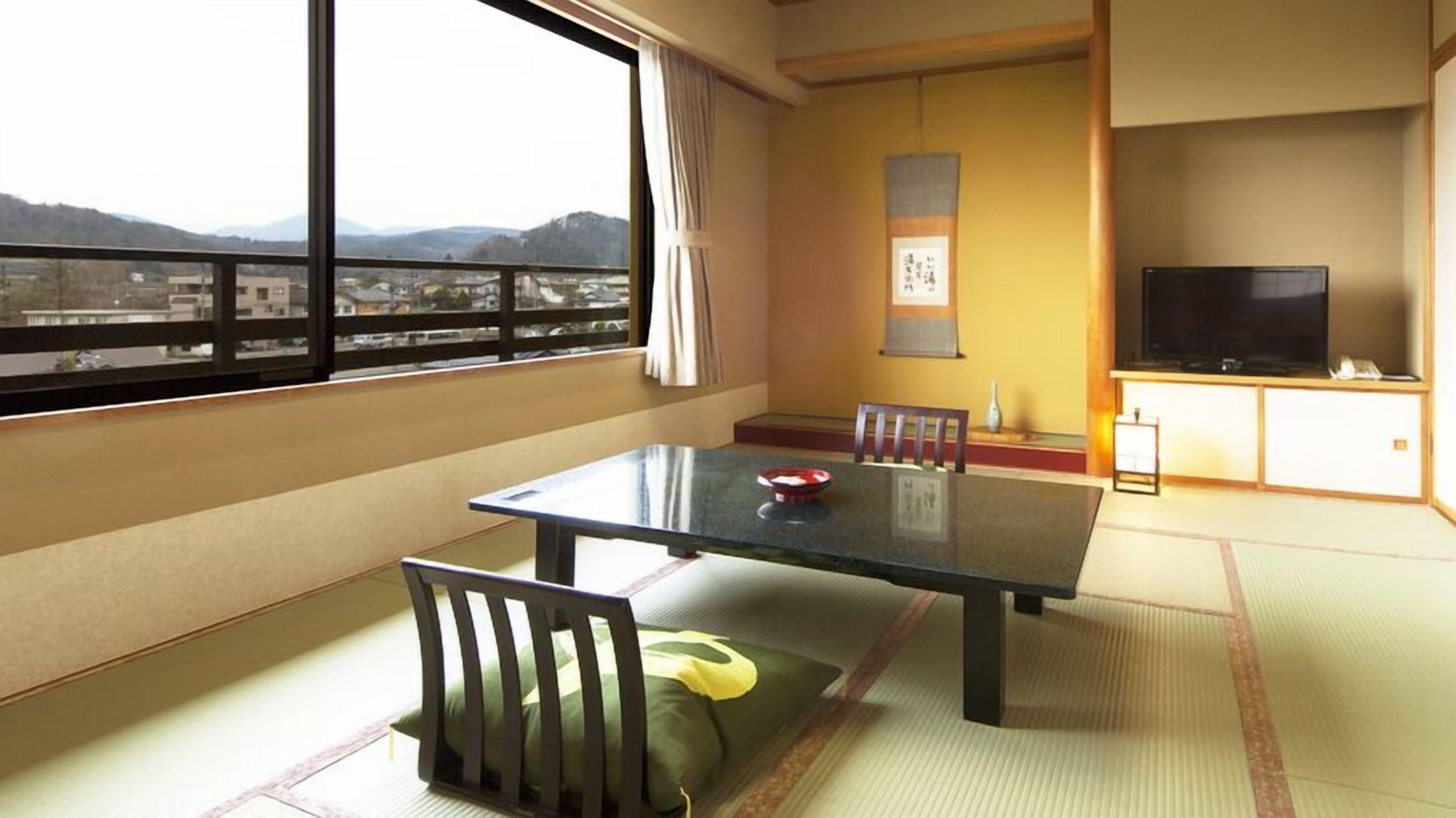A room with tatami flooring, a low table with a cushion, a television, and a view of a cityscape through large windows.