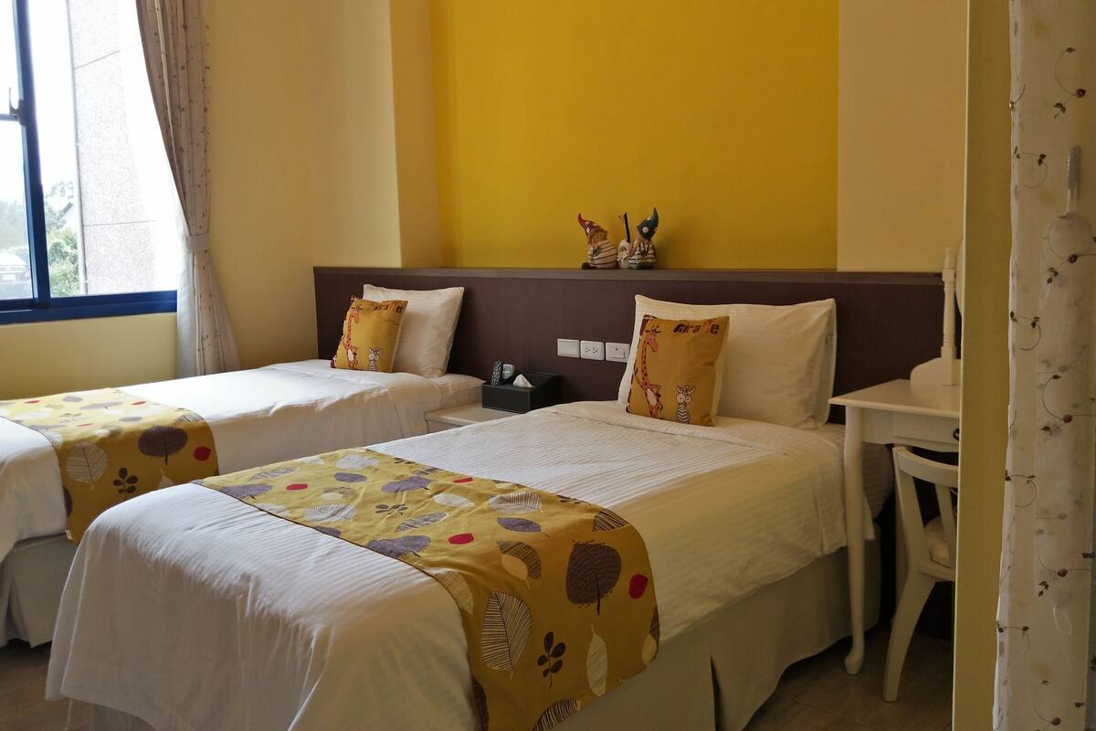 A hotel room with two beds, a nightstand, a small table, and a window with curtains.
