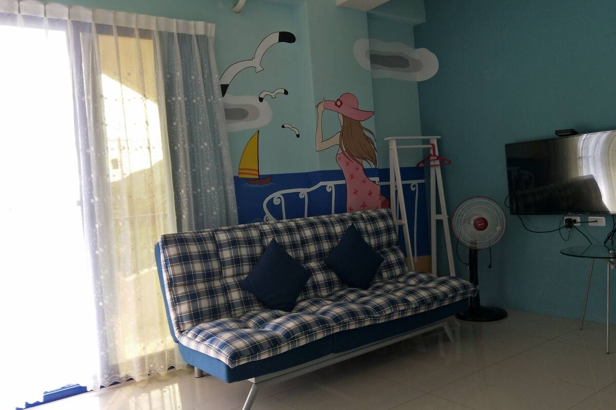 A living room with a sofa, a TV, a fan, and a wall with a sailboat mural.