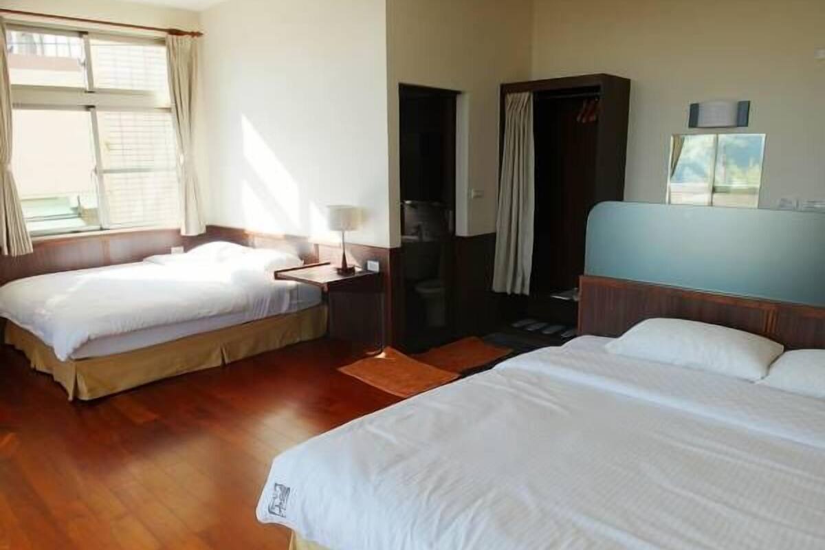 A hotel room with two single beds, a wooden floor, a desk with a lamp, a wardrobe, and a window with curtains.