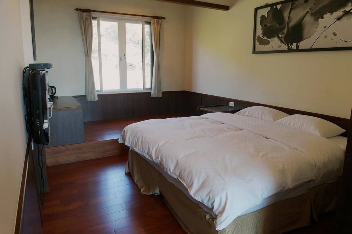 A bedroom with a large bed, a desk, a chair, a window with curtains, and a painting on the wall.