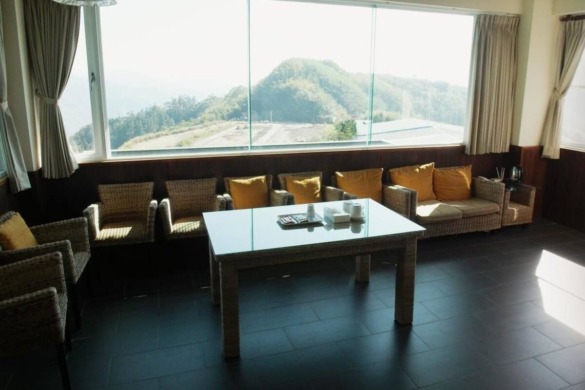 A room with wicker furniture, a glass-top table, and a large window offering a view of a hilly landscape.