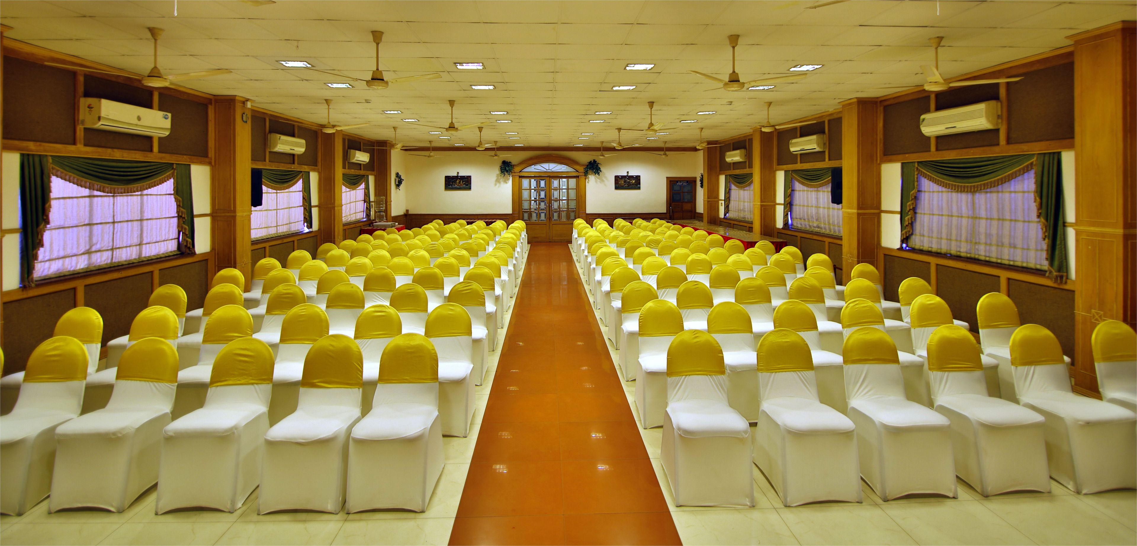 Un salón de conferencias con filas de sillas blancas adornadas con cintas amarillas, paneles de madera y ventiladores de techo.