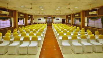Un salón de conferencias con filas de sillas blancas adornadas con cintas amarillas, paneles de madera y ventiladores de techo.
